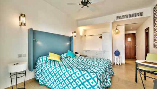 Et hotelværelse med en dobbeltseng, blå sengegavl, farverige puder og et mønstret tæppe. Detaljer inkluderer møbler og dekorative elementer. (AI-genereret tekst)