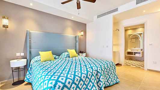 Et hotelværelse med en seng med et blåt og hvidt mønstret sengelinned og gule puder. I baggrunden ses et badeværelse. (AI-genereret tekst)