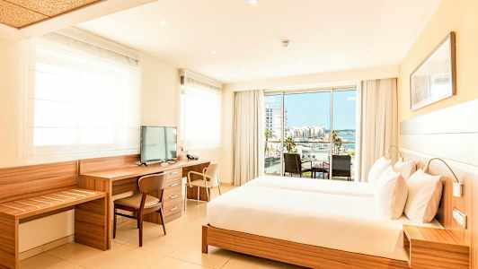 Et lyst hotelværelse med en stor seng, skrivebord og tv samt en balkon med udsigt over en by og havet. (AI-genereret tekst)