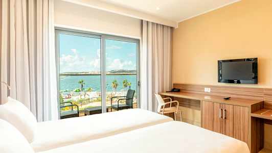 Et hotelværelse med to senge og udsigt over havet gennem en glasdør. (AI-genereret tekst)