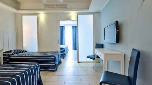 To enkeltsenge med stribede blå sengetæpper, et hvidt skrivebord med to blå stole og et vægmonteret TV samt en glasdør til værelset bagved. (AI-genereret tekst)