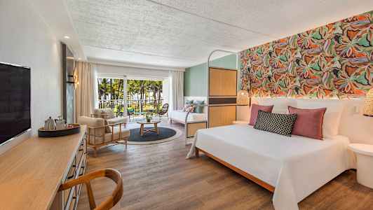 Hotelværelse med dobbeltseng, siddeområde, skrivebord og altan med udsigt over palmer og have. (AI-genereret tekst)