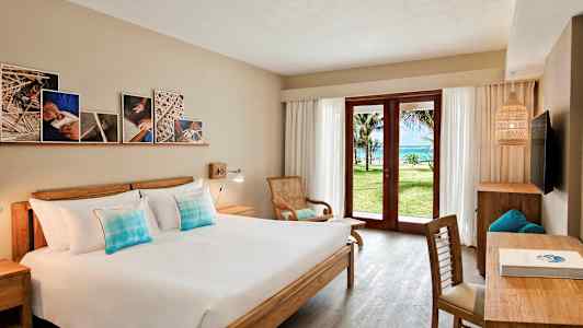 Et hotelværelse med en dobbeltseng, træmøbler og en glasdør, der åbner ud til en grøn have og udsigt til havet. (AI-genereret tekst)