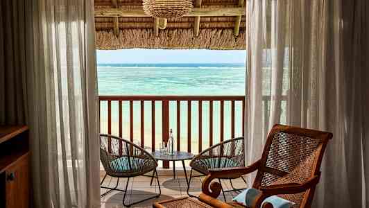 Udsigt over havet fra et værelse med en overdækket altan. Mellem gennemsigtige gardiner ses to havestole ved et bord. (AI-genereret tekst)