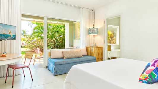 Et lyst hotelværelse med seng, siddeområde og bord. Udsigt til haven kan ses gennem glasdørene. (AI-genereret tekst)