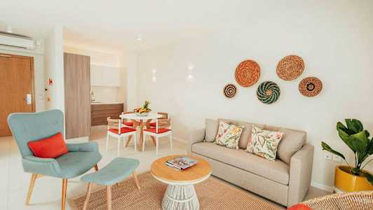 En lys og moderne indrettet stue med beige sofa, grøn lænestol og et rundt sofabord af træ. Køkkenet er synligt i baggrunden. (AI-genereret tekst)