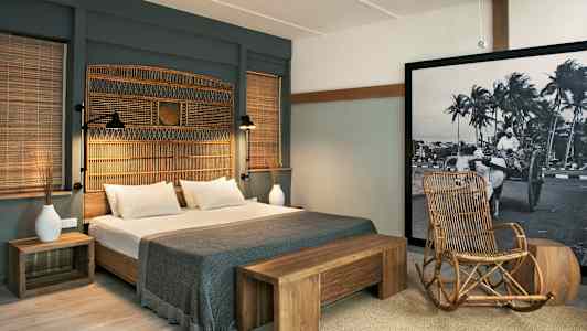 Et hotelværelse med en dobbeltseng, trædetaljer og en stol af rattan. Et stort billede med sort-hvidt motiv pryder væggen. (AI-genereret tekst)