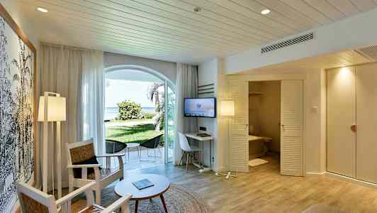 Et hotelværelse med siddepladser, et bord og et TV. En dør fører til et badeværelse, og der er vinduer, der vender ud mod en grøn have og stranden. (AI-genereret tekst)