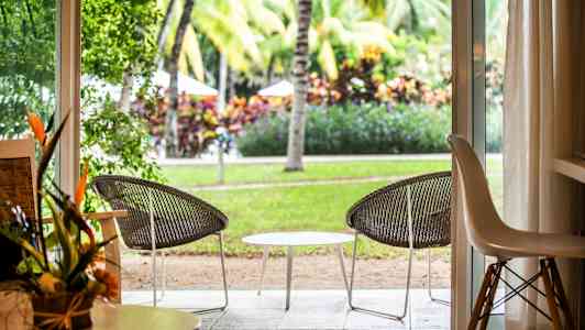 Udsigt fra et værelse gennem en åben dør til en terrasse med to stole og et bord, omgivet af grønne områder og palmer. (AI-genereret tekst)
