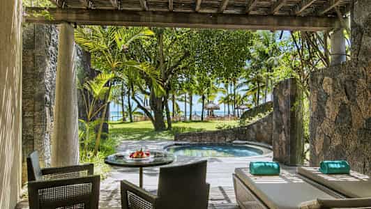 En terrasse med siddepladser og liggestole ved siden af en lille pool, omgivet af planter og med udsigt mod stranden og palmetræer. (AI-genereret tekst)