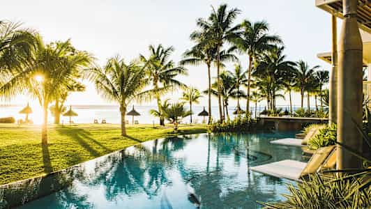 Udsigt over en pool med liggestole omgivet af palmer og udsigt til stranden ved havet. (AI-genereret tekst)