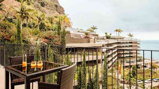 Billede af en altan med rotanmøbler, med et bord der har to glas og en karaffel. Udsigt mod beplantning, bygninger og havet. (AI-genereret tekst)