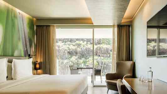 Et hotelværelse med en dobbeltseng, siddegruppe, et bord med stole samt en stor glasdør, der åbner op til en balkon med udsigt over natur og klipper. (AI-genereret tekst)