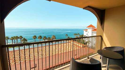 Udsigt fra en balkon med to stole og et bord, mod havet og palmer. (AI-genereret tekst)