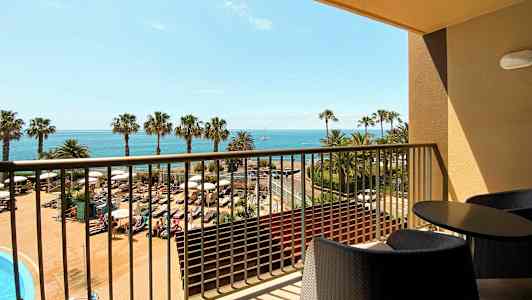 Udsigt fra en altan med møbler over et poolområde, palmer og havet i baggrunden. (AI-genereret tekst)