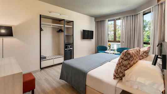 Et hotelværelse med dobbeltseng, to blå stole ved et bord foran vinduer samt garderobe med åbne hylder. (AI-genereret tekst)