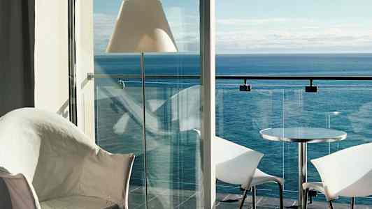 Udsigt fra et værelse mod havet med en balkon med glasrækværk, bord og stole. (AI-genereret tekst)