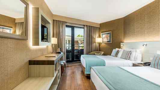 Et hotelværelse med to enkeltsenge, en skrivebordsflade, et vægmonteret TV og et vindue, der fører ud til en altan. (AI-genereret tekst)