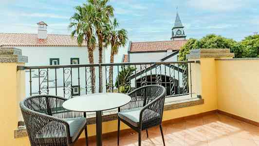 Billedet viser en altan med to stole og et bord. Udsigt til et hvidt hus med rødt tag og palmer. (AI-genereret tekst)