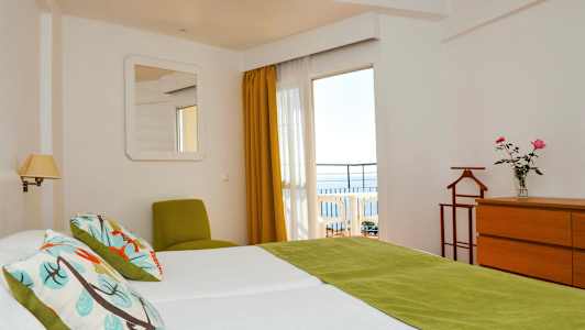 Et hotelværelse med to senge, grønne detaljer og udsigt til en altan med havet i baggrunden. (AI-genereret tekst)