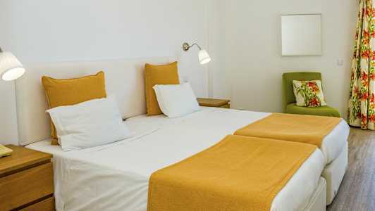 Et hotelværelse med to enkeltsenge og gult sengetøj. En grøn lænestol med blomstrede puder og gardiner ses i baggrunden. (AI-genereret tekst)