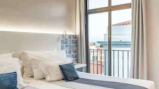 Et hotelværelse med en dobbeltseng, hvide sengelagner og blå puder, ved siden af et vindue med udsigt. (AI-genereret tekst)