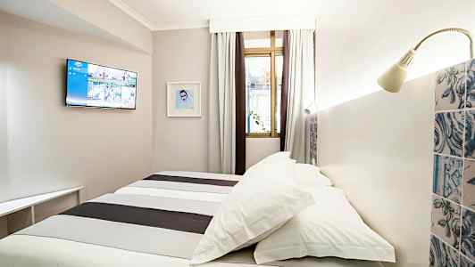 Et hotelværelse med en seng, vægmonteret tv og et vindue med gardiner. (AI-genereret tekst)