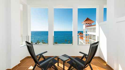 To sorte stole og et bord på en terrasse med udsigt over havet og en hvid bygning med røde tage. (AI-genereret tekst)