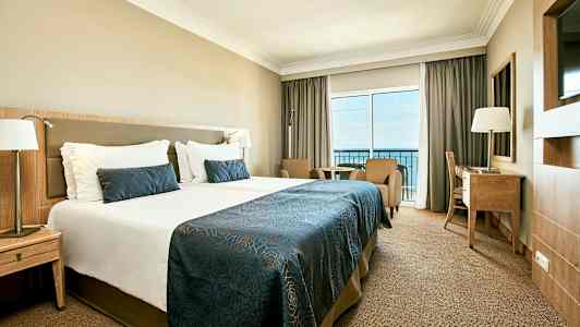 Billede af et hotelværelse med en stor seng, to lænestole, et skrivebord og en balkon med udsigt over havet. (AI-genereret tekst)