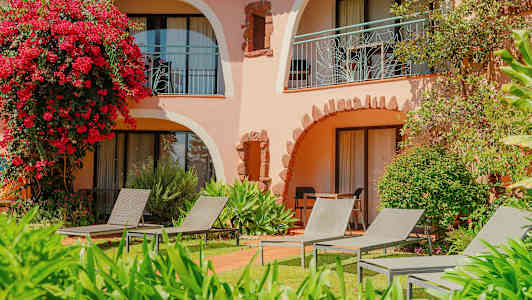 En bygning med buede altaner omgivet af grønt og blomster, med liggestole placeret på græsplænen. (AI-genereret tekst)