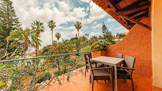 En terrasse med bord og stole omgivet af grønne planter og palmer. (AI-genereret tekst)