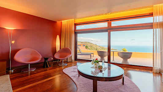 Et rum med røde vægge, to stole, et bord og store vinduer der vender ud mod en terrasse med havudsigt. (AI-genereret tekst)