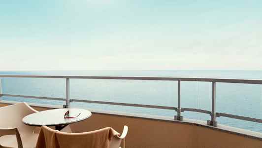 Udsigt fra en altan med bord, stole og glasrækværk, havet ses i baggrunden. (AI-genereret tekst)