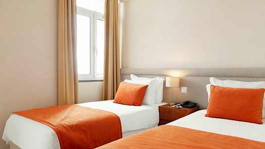 Et hotelværelse med to enkeltsenge ved siden af hinanden, med hvide lagner og orange tæpper og pyntepuder. (AI-genereret tekst)