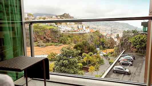 Udsigt fra en altan med møbler mod et bjergrigt område med bygninger og parkerede biler. (AI-genereret tekst)