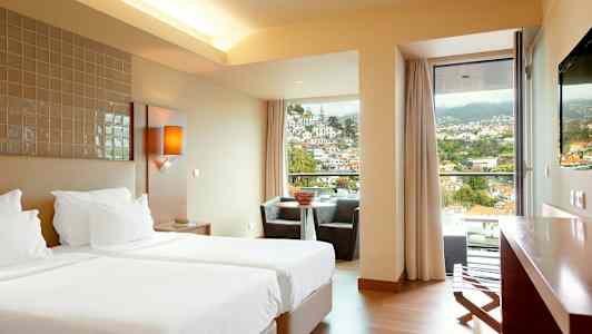 Et hotelværelse med to enkeltsenge, en siddegruppe ved et vindue med udsigt over en by i et bjergområde. (AI-genereret tekst)