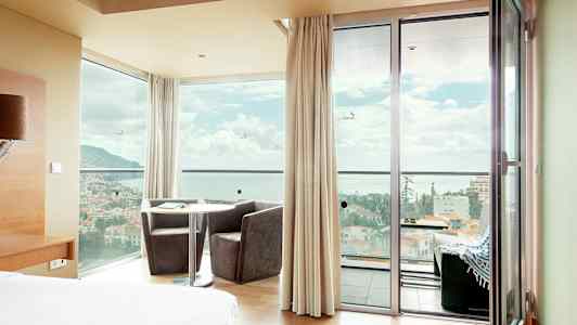 Hotelværelse med panoramisk udsigt gennem vinduer og døre over en by foran havet. (AI-genereret tekst)