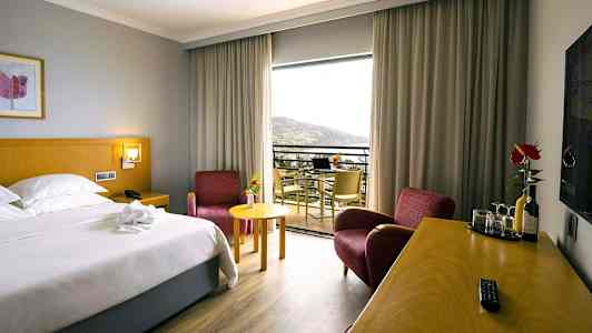 Hotelværelse med en dobbeltseng, to røde lænestole, et skrivebord med drikkevarer og glas og en balkon med udsigt. (AI-genereret tekst)