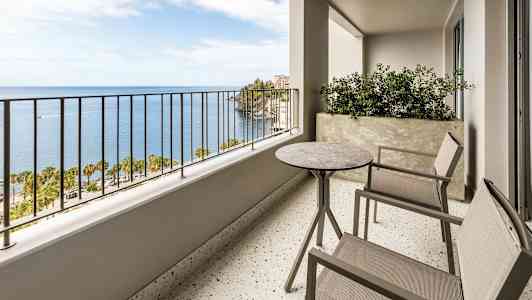 En balkon med to stole og et bord samt en udsigt over havet og palmer. (AI-genereret tekst)