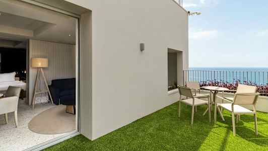 En terrasse med kunstgræs, bord og stole samt havudsigt, forbundet til et moderne rum med indretning. (AI-genereret tekst)