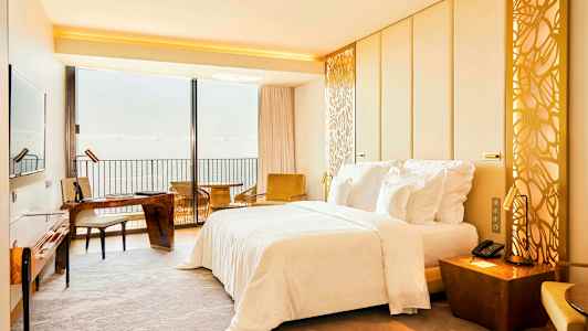 Hotelværelse med en dobbeltseng, skrivebord og udsigt over havet gennem en stor vinduesvæg. (AI-genereret tekst)