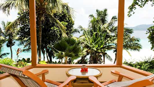 Træterrasse med to stole og et bord med udsigt over tropiske palmer og blåt hav. (AI-genereret tekst)