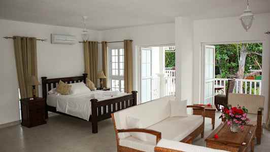 Et lyst hotelværelse med en seng, sofa og åbne døre til en balkon med udsigt over naturen. (AI-genereret tekst)