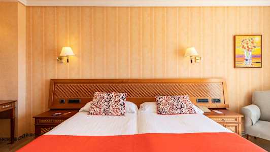 Et hotelværelse med en dobbeltseng med hvidt og orange sengetøj og dekorative puder. Sengegavlen er af træ med et indlæg af rutemønster. (AI-genereret tekst)