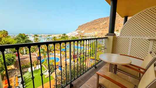 Udsigt fra altan over poolområde med frodighed, palmer og en bjergside i baggrunden. (AI-genereret tekst)