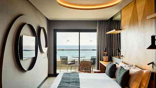 Et moderne hotelværelse med seng, spejle på væggen, skrivebord og en balkon med udsigt over havet. (AI-genereret tekst)