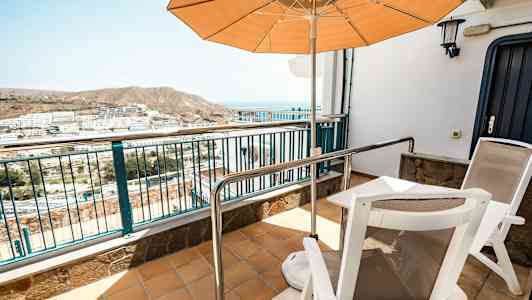 En terrasse med to stole, et bord og en parasol med udsigt over et boligområde og bjerge i baggrunden. (AI-genereret tekst)