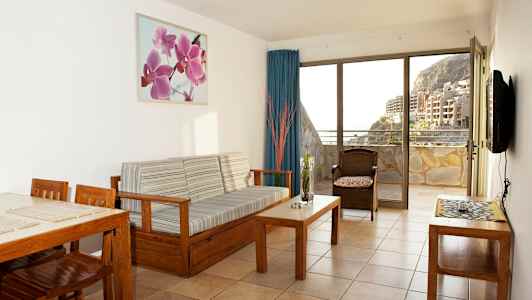 En stue med træindretning og udsigt til en altan samt bygninger og bjerge. (AI-genereret tekst)