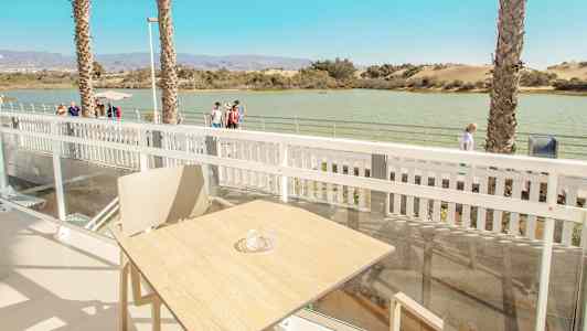 Udsigt fra en terrasse med bord og stol, med vand og gangsti i baggrunden. (AI-genereret tekst)