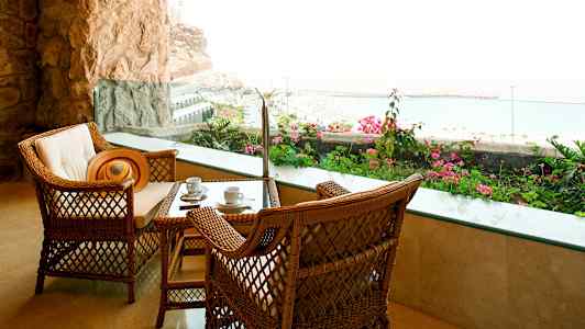 To flettede stole og et bord med kaffekopper, på en terrasse med udsigt til havet og blomsterpragt. (AI-genereret tekst)